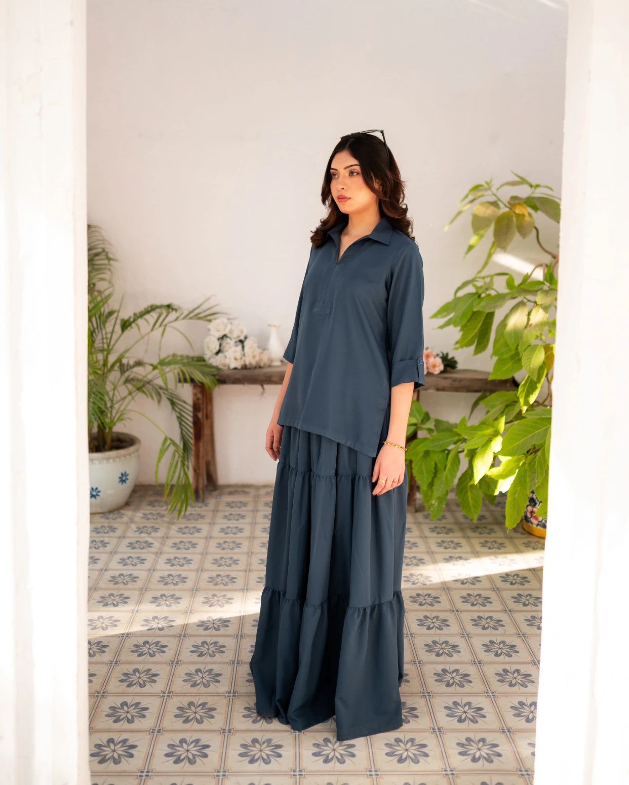 Woman wearing a steel blue skirt and shirt standing in a room with plants and a tiled floor.