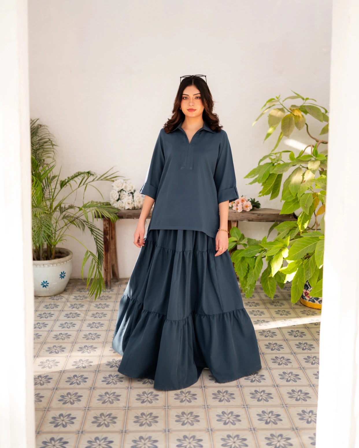 Woman wearing a steel blue shirt with a long skirt in a room with plants and a tiled floor.
