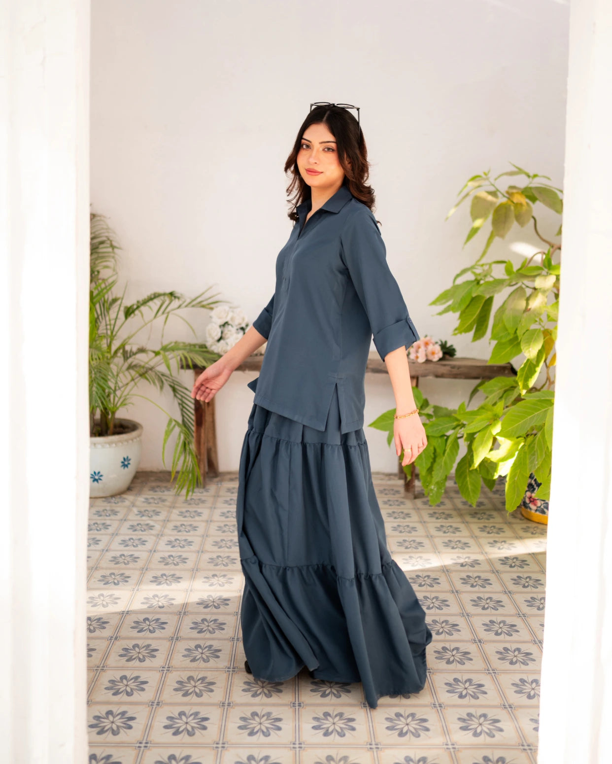 Woman wearing a steel blue skirt and shirt standing in a room with plants and a tiled floor.