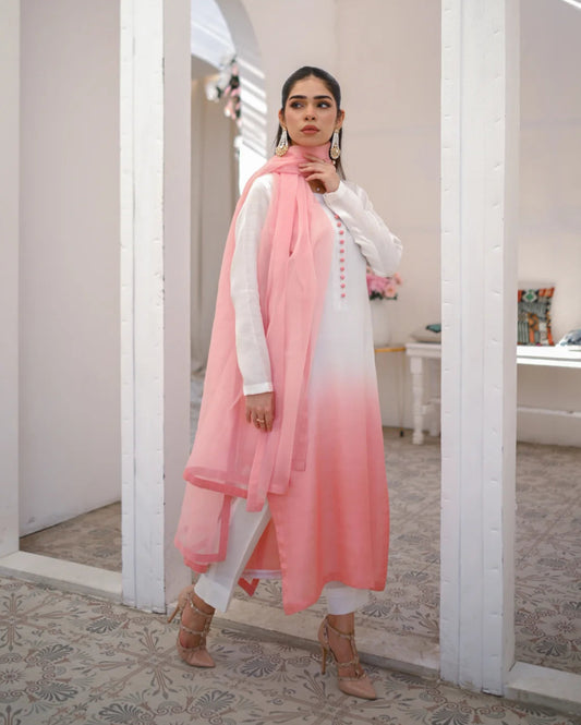 A woman wearing a three piece rawsilk dress with an ombre shirt, trousers, and an organza dupatta in pink and white colors.