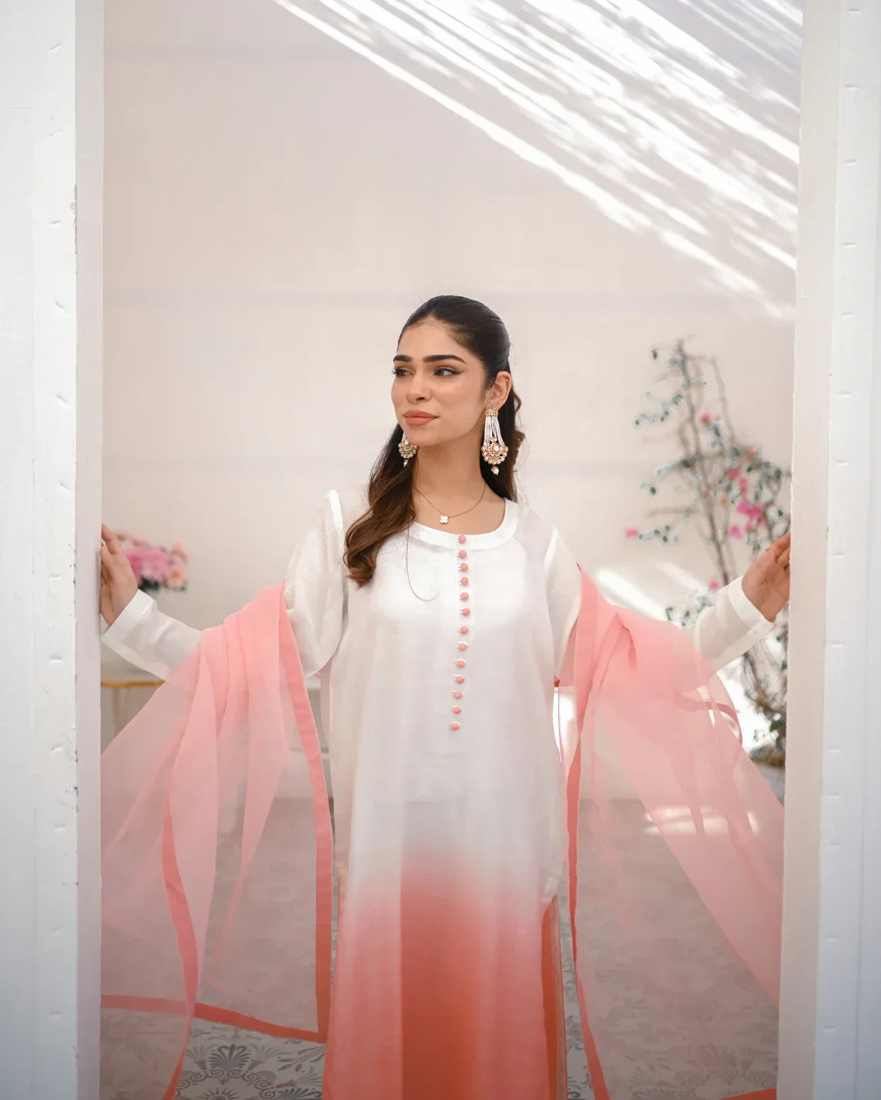 A woman wearing a three piece rawsilk dress with an ombre shirt, trousers, and an organza dupatta in pink and white colors.