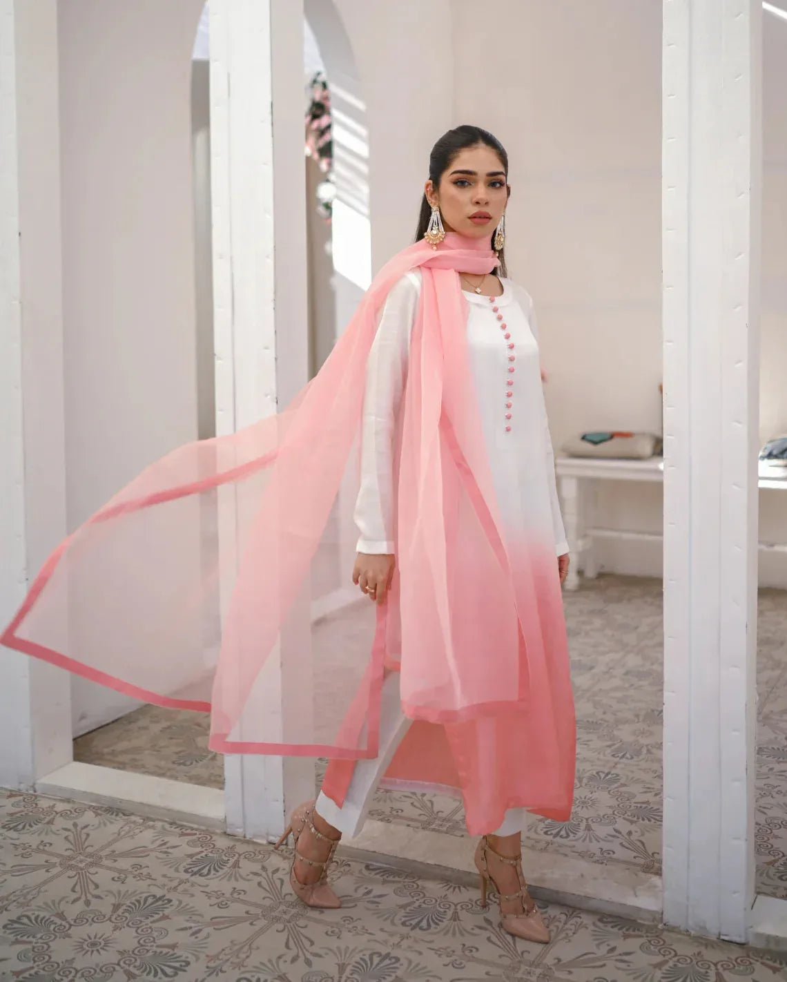 A woman wearing a three piece rawsilk dress with an ombre shirt, trousers, and an organza dupatta in pink and white colors.