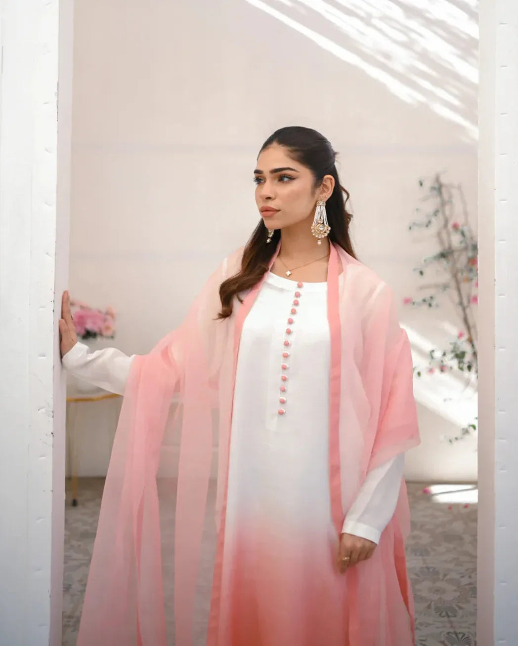 A woman wearing a three piece rawsilk dress with an ombre shirt, trousers, and an organza dupatta in pink and white colors.