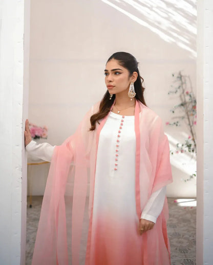 A woman wearing a three piece rawsilk dress with an ombre shirt, trousers, and an organza dupatta in pink and white colors.