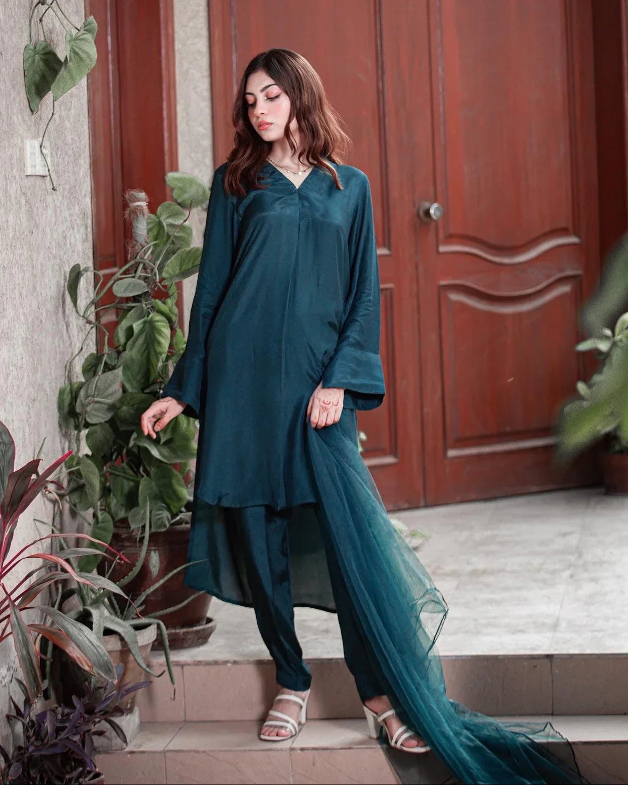 Woman in a teal blue dress standing in front of a wooden door with plants around.