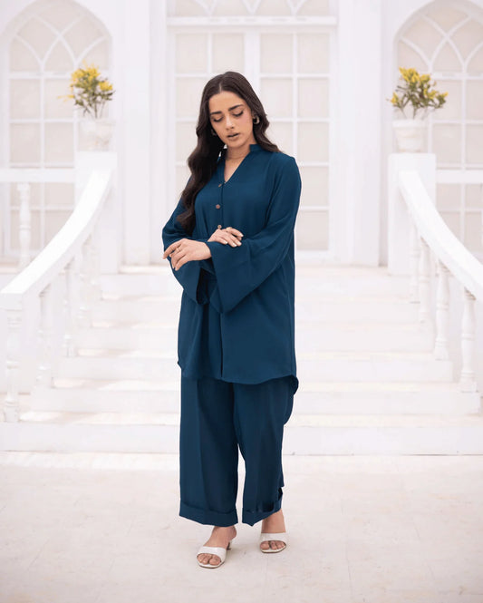 A woman modeling a teal blue two-co ord set with wide bell sleeves and matching wide-leg pants, standing in a bright white architectural setting with stairs and arches.