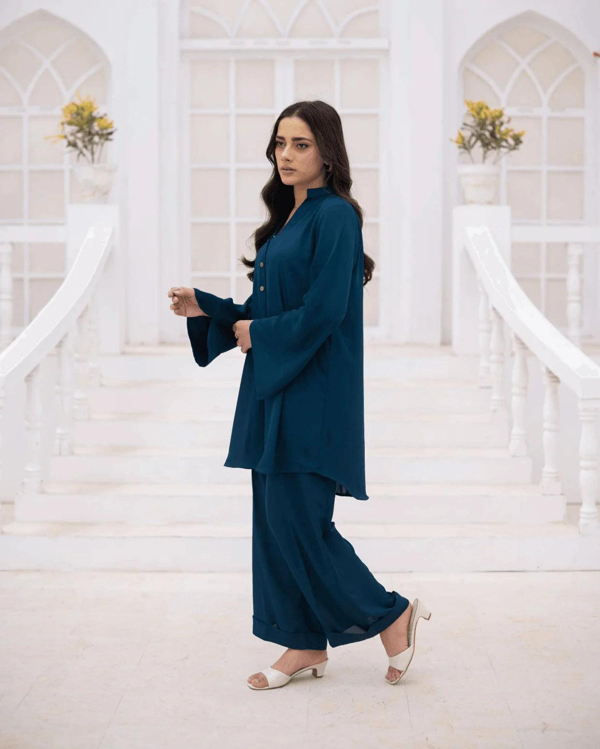 A woman models a teal blue co ord set consisting of a long-sleeve button-up top, a matching jacket, and wide-leg trousers set against a bright white architectural background with white stairs and arches.