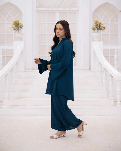 A woman models a teal blue co ord set consisting of a long-sleeve button-up top, a matching jacket, and wide-leg trousers set against a bright white architectural background with white stairs and arches.