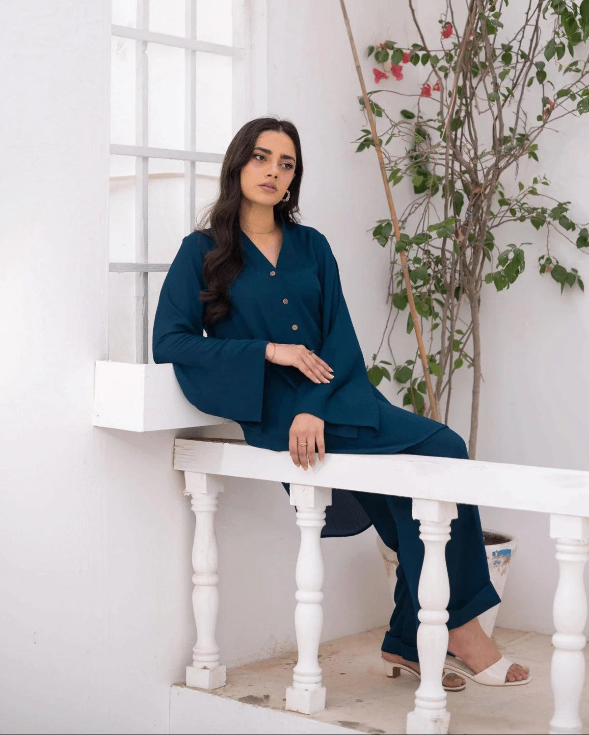 A woman wearing a teal blue co ord set, sitting on a white wooden balcony railing against a white wall with a potted plant.