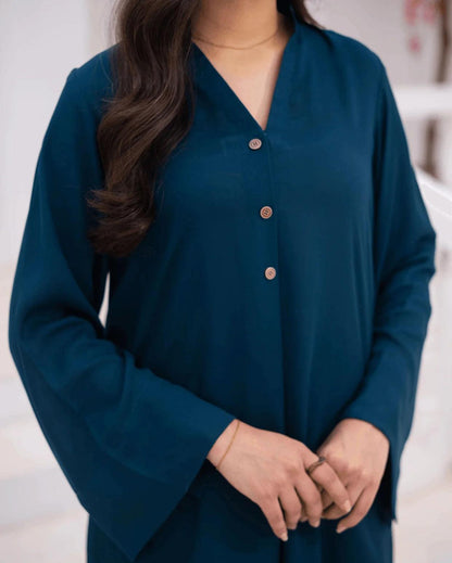 A woman wearing a teal blue long-sleeve button-up top with a V-neck and metallic buttons, shown from the chest down against a softly blurred light background.