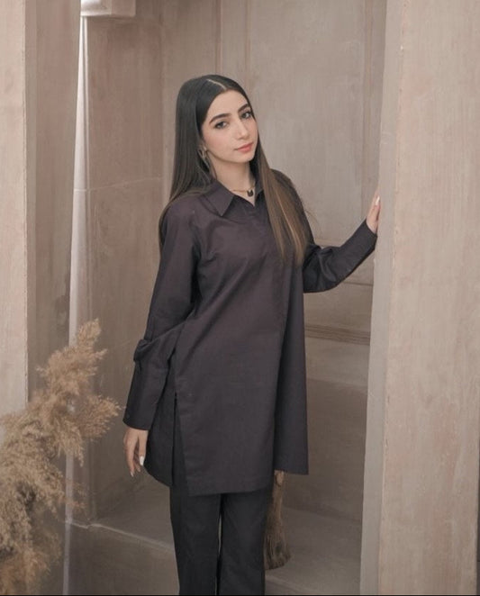 A woman in a stylish dark brown outfit stands in a minimalist room with decorative vases and pampas grass. The ambiance is calm and elegant.