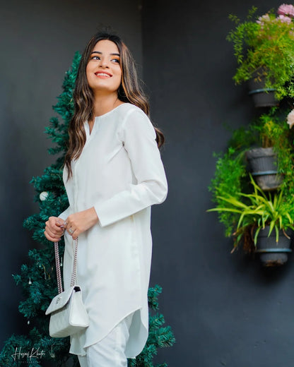 woman wearing white dress stands beside a decorated tree and hanging plants. 