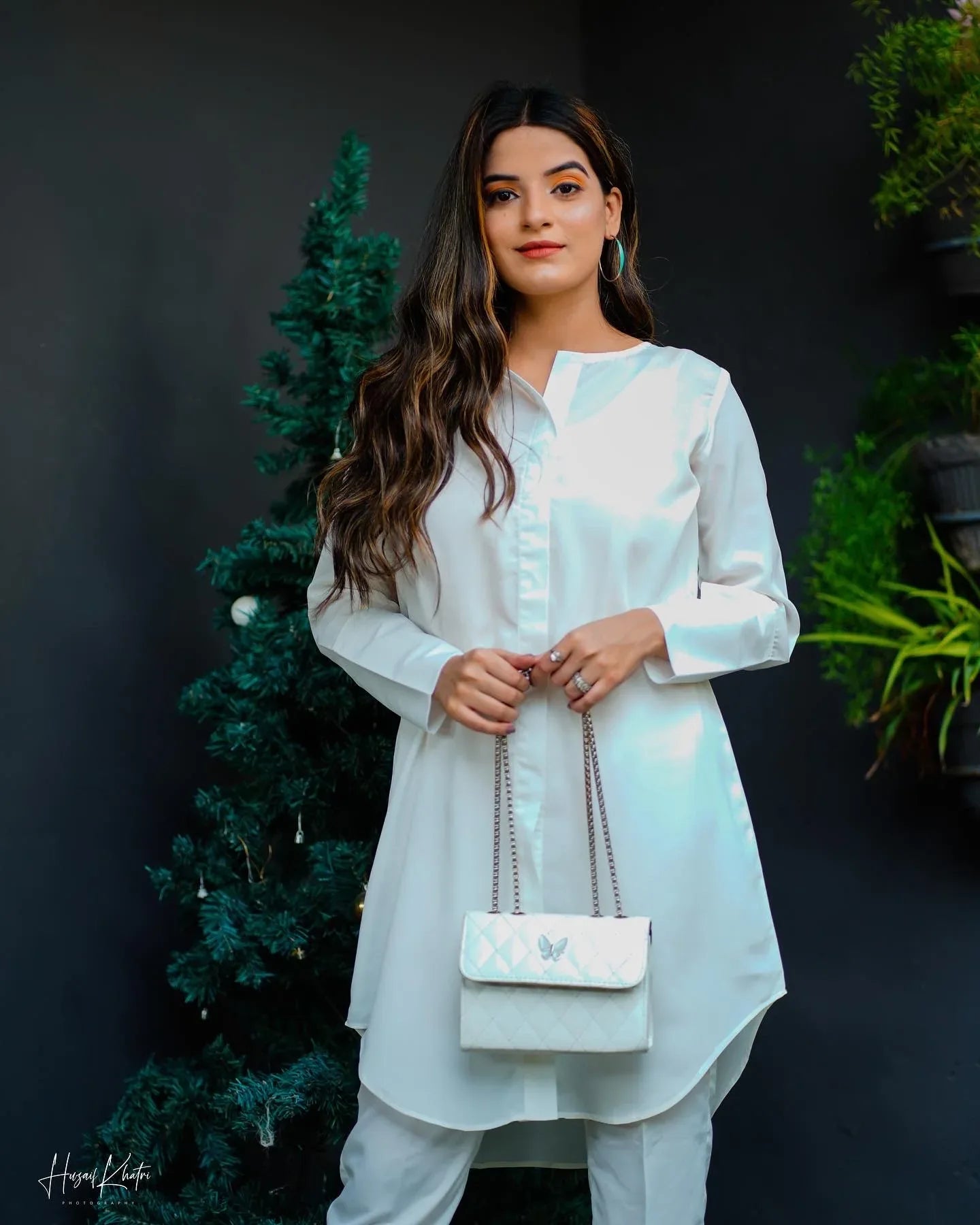 woman wearing a white dress stands beside a decorated tree and hanging plants. 