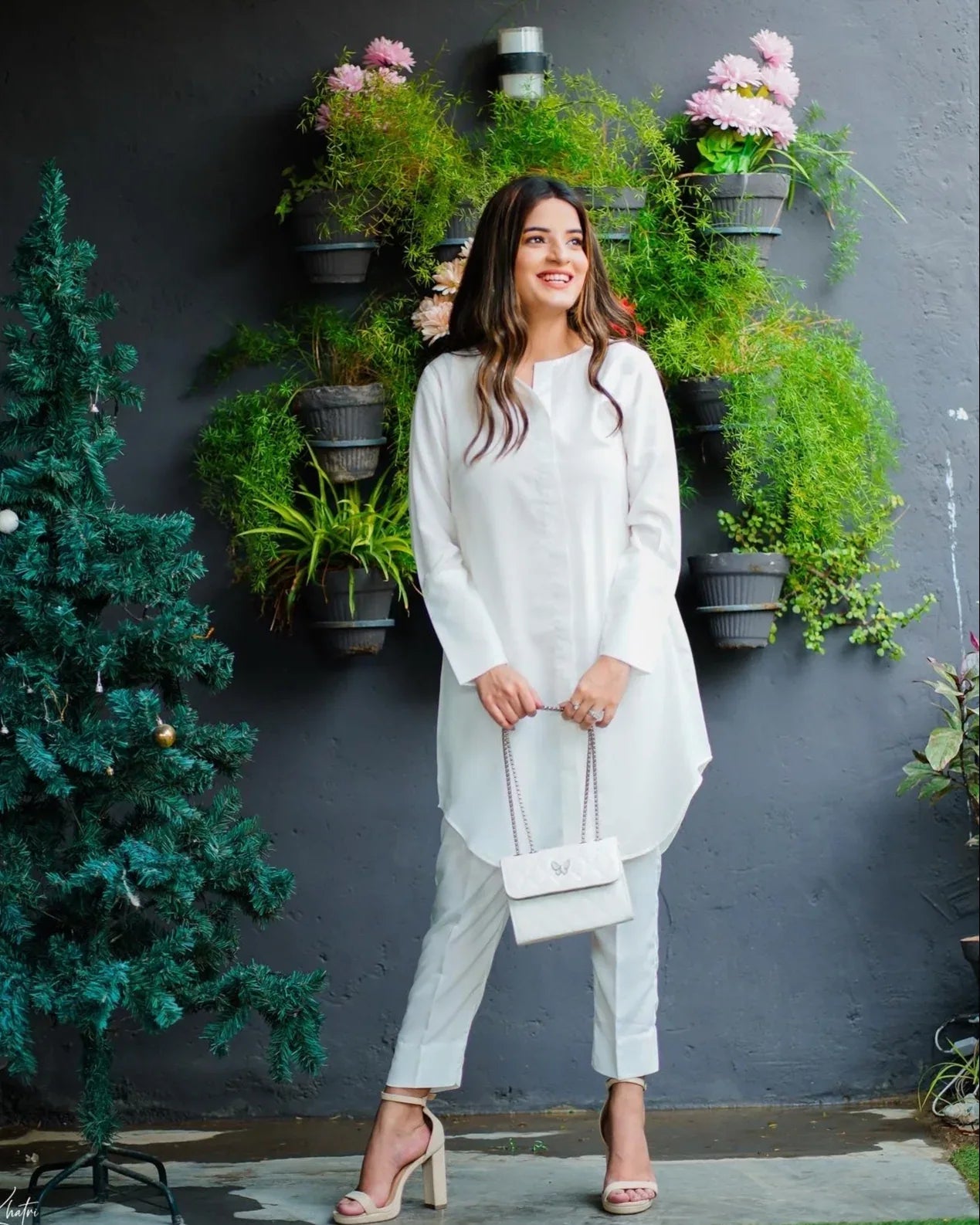 woman wearing a white dress stands beside a decorated tree and hanging plants. 