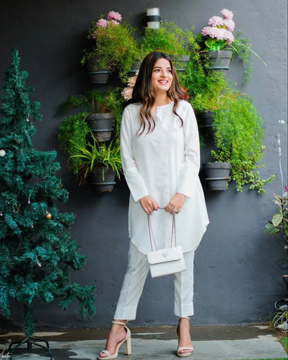 woman wearing a white dress stands beside a decorated tree and hanging plants. 