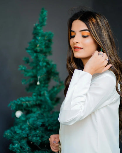 woman wearing a white dress stands beside a decorated tree and hanging plants. 