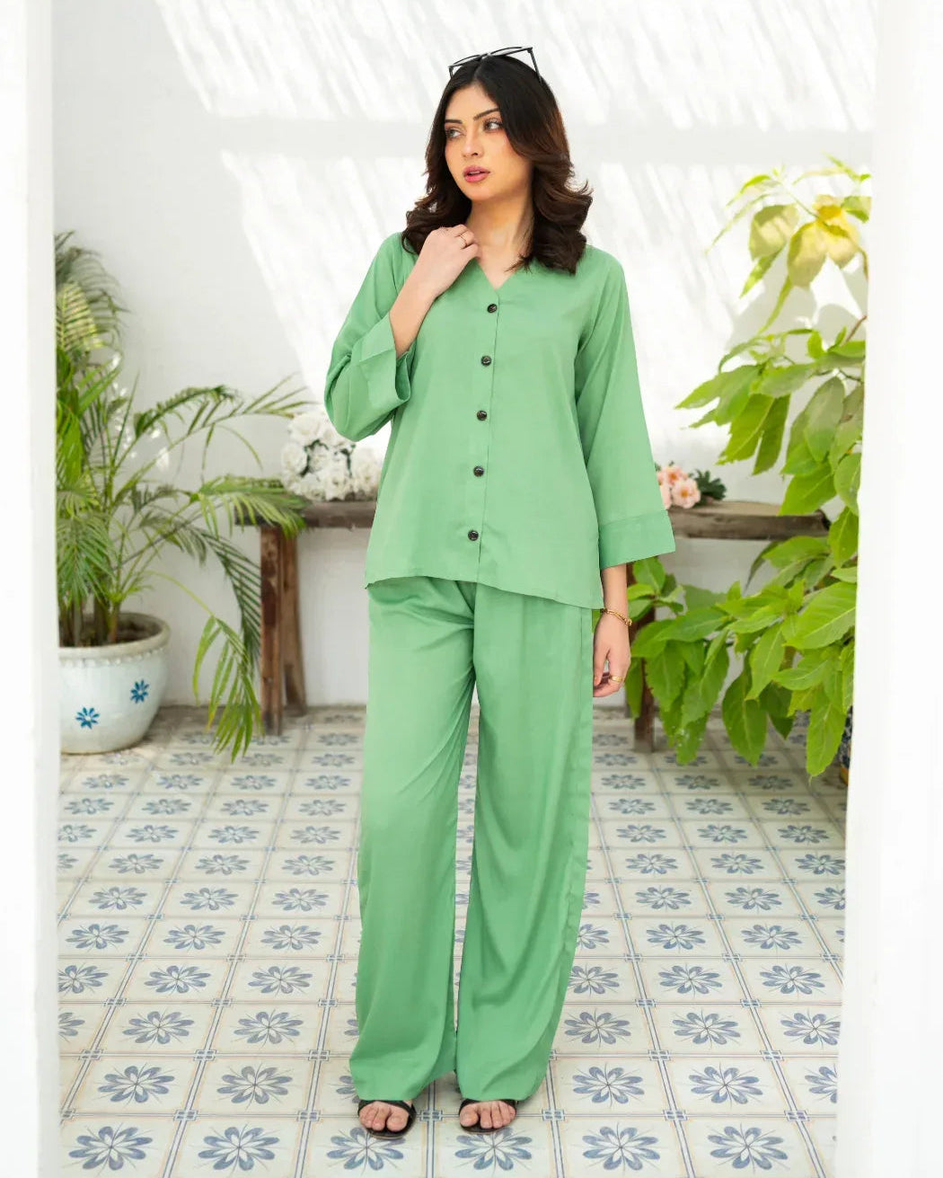 Woman wearing a green outfit standing in a room with plants and patterned tiles.