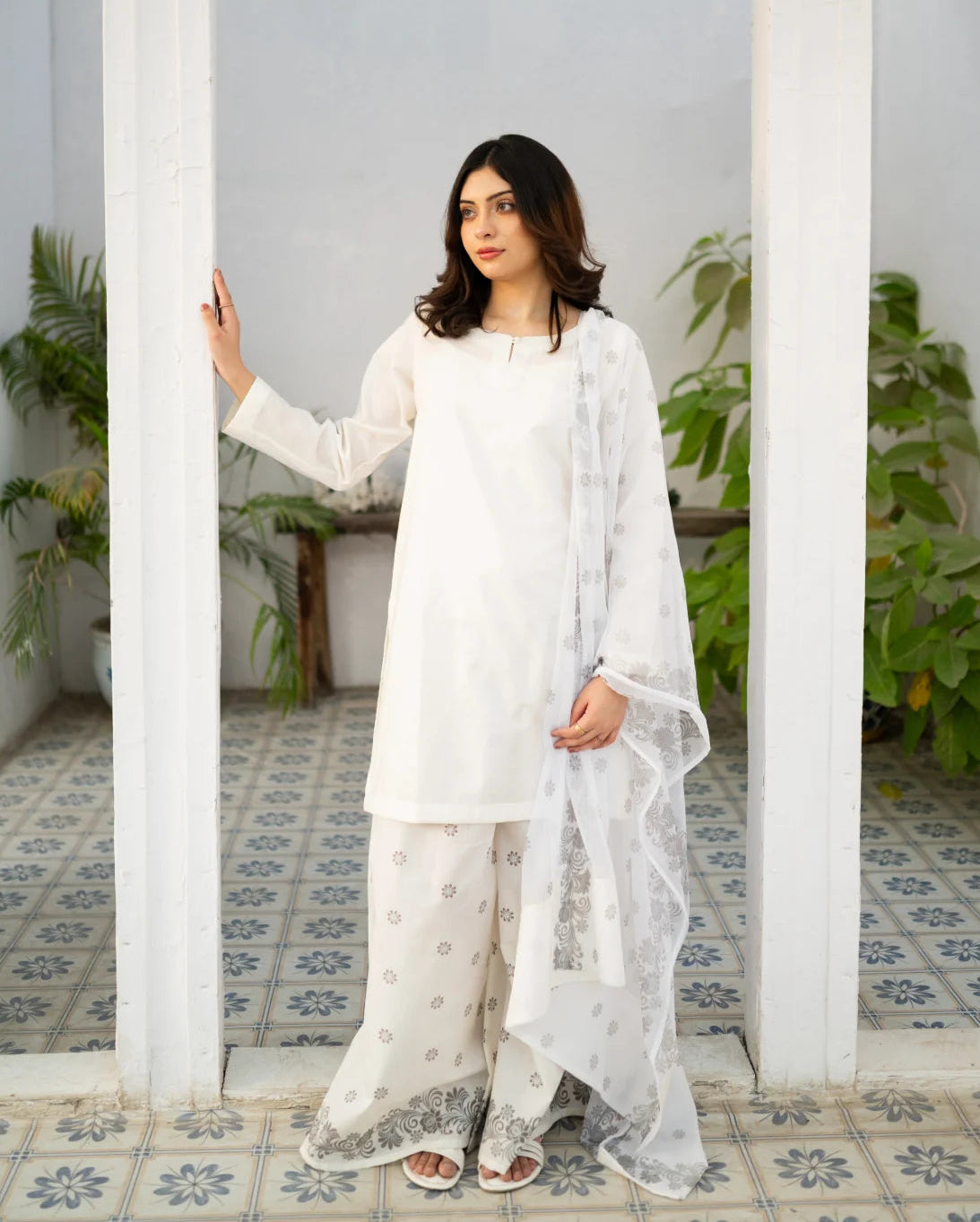 Woman in a white dress, a traditional pakistani kurta set standing in a room with white walls and tiled floor.